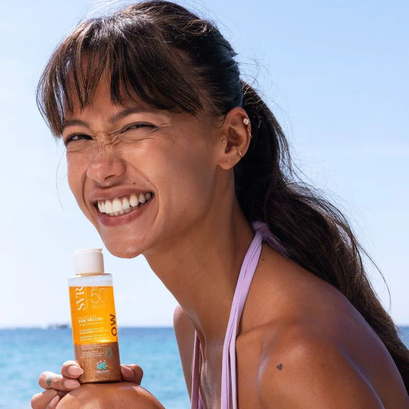 Gros plan d'une femme souriante tenant le SVR Sun Secure Eau Solaire Glow SPF50+.