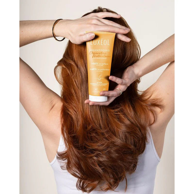 Femme aux longs cheveux roux vue de dos tenant le tube de Shampooing Réparateur Moléculaire Luxéol, 