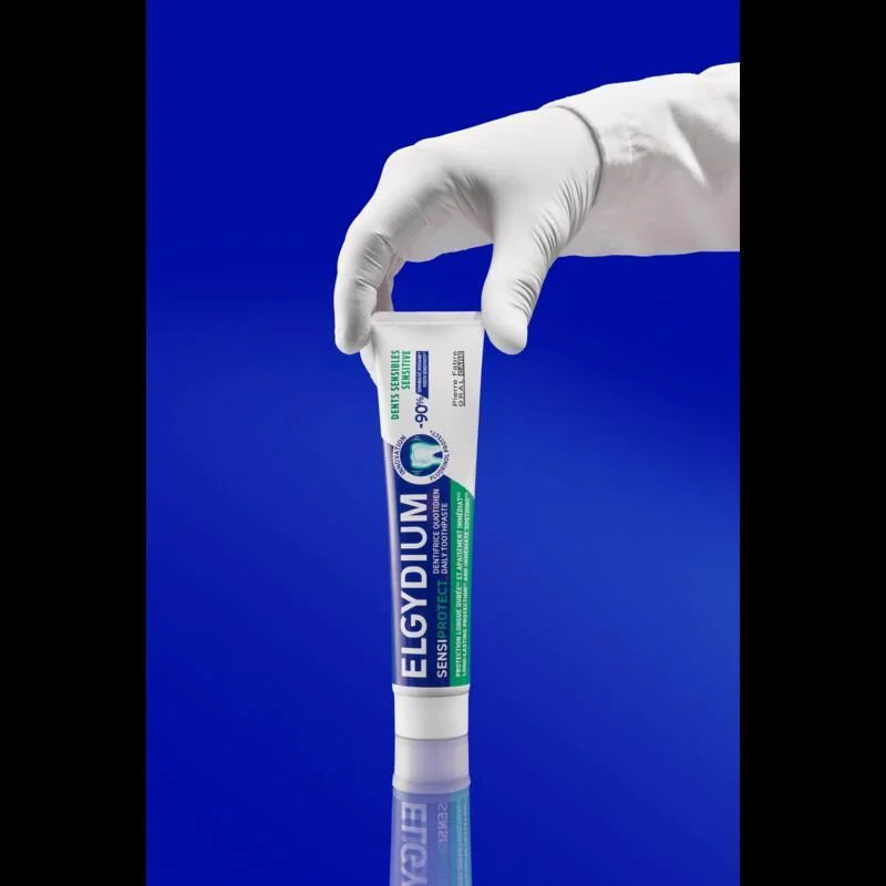 Tube et boîte de dentifrice Elgydium Sensiprotect présentés ensemble sur surface bleue avec reflet, 