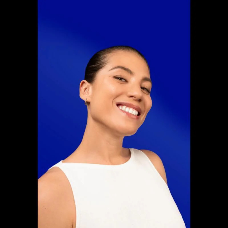 Main gantée de blanc tenant le tube Elgydium Sensiprotect sur fond bleu, femme souriante montrant se